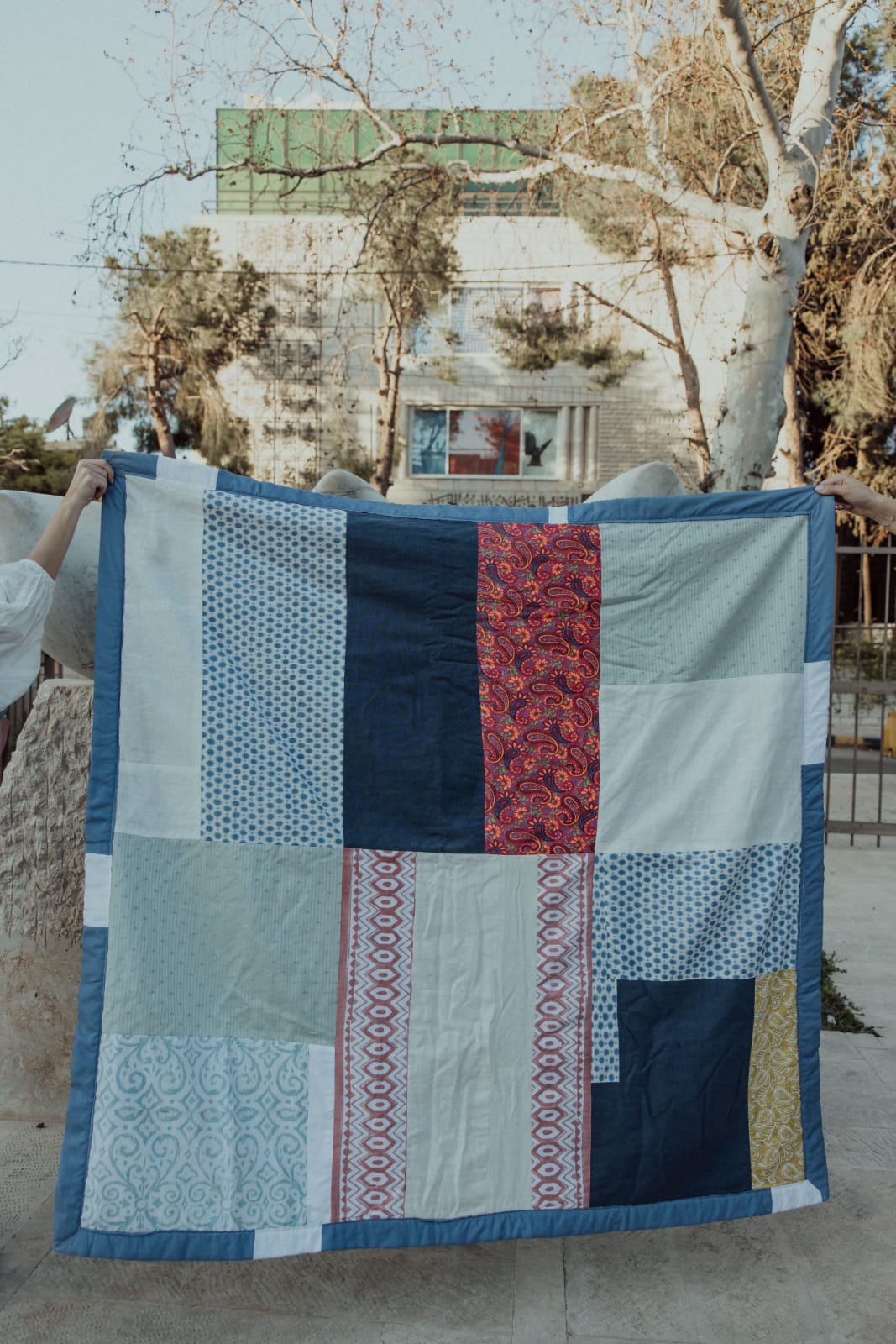 Large patchwork quilt in blues, creams and red paisley, held up on a street